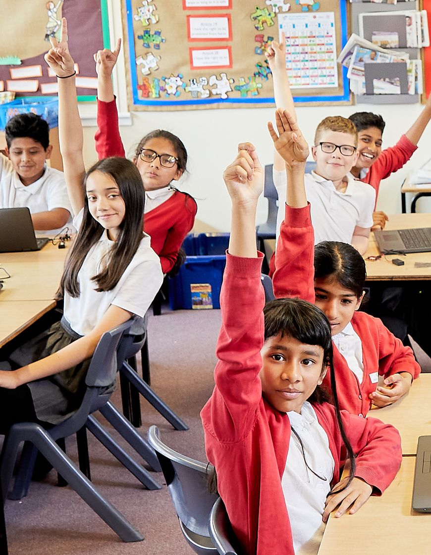 Seven children in school uniform, sitting in their classroom, with their hands raised